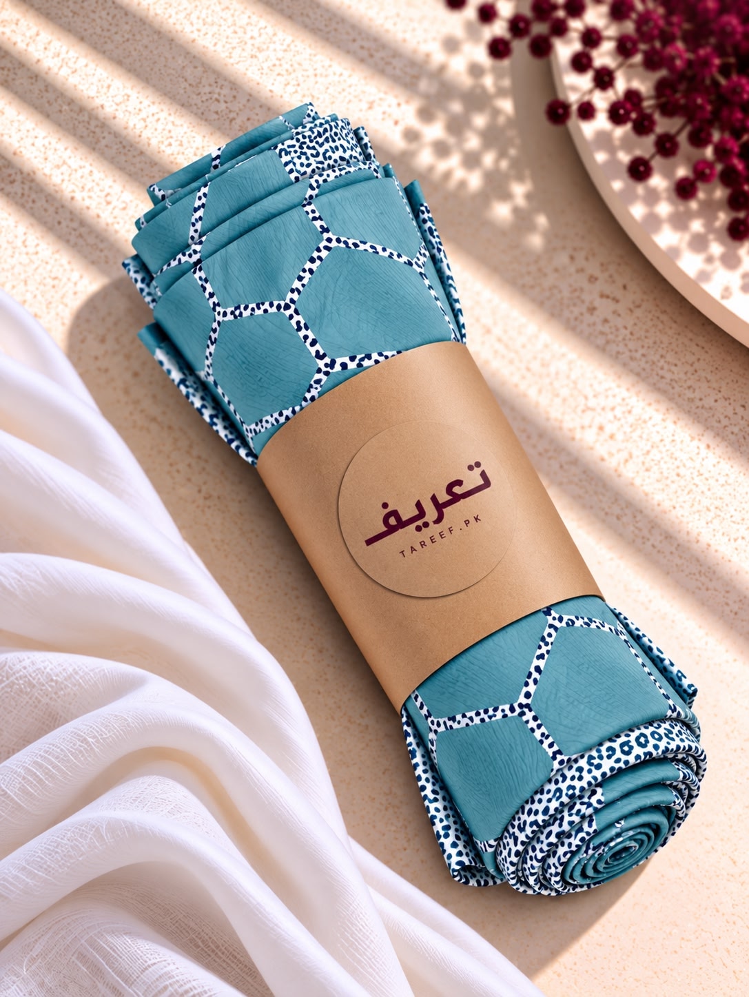 A rolled teal hijab with a white geometric lattice pattern and a white border, wrapped in a brown paper band with the brand name 'TAREEF' .