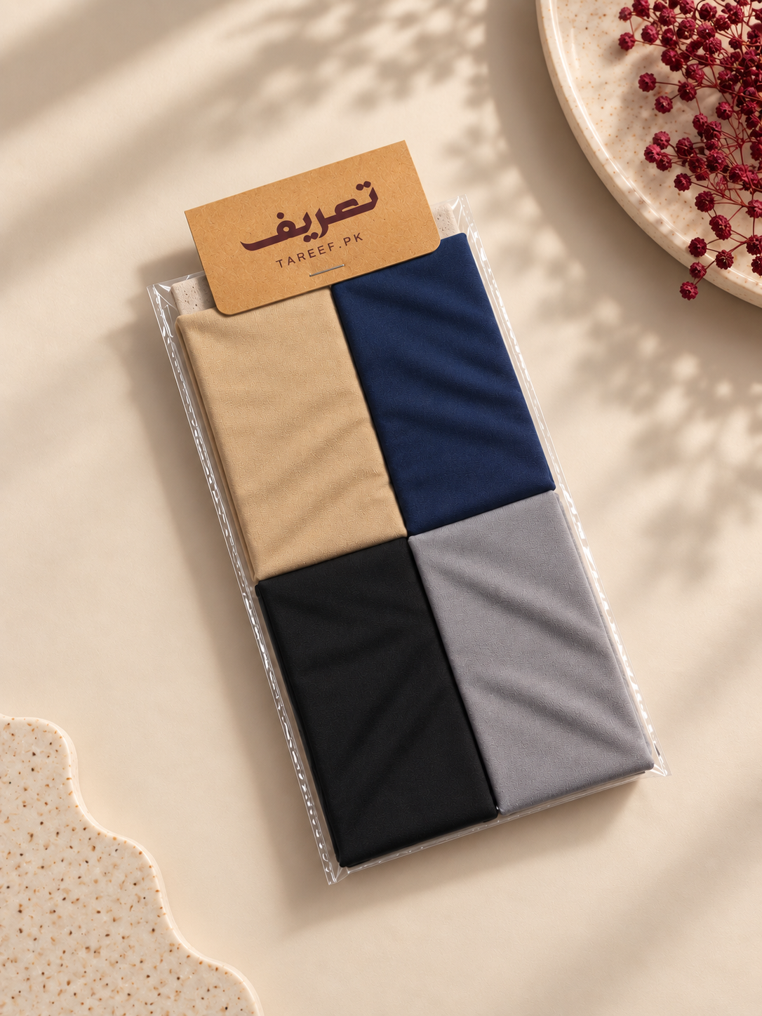 A packaged hijab tube in beige, navy, black, and light gray, with a kraft paper label reading 'TAREEF' on a warm beige styled background with decorative dried red berries and textured props.