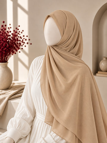 A beige draped hijab styled on a white mannequin wearing a white long-sleeve dress, set in a warm, sunlit interior with a vase of red flowers on the left.