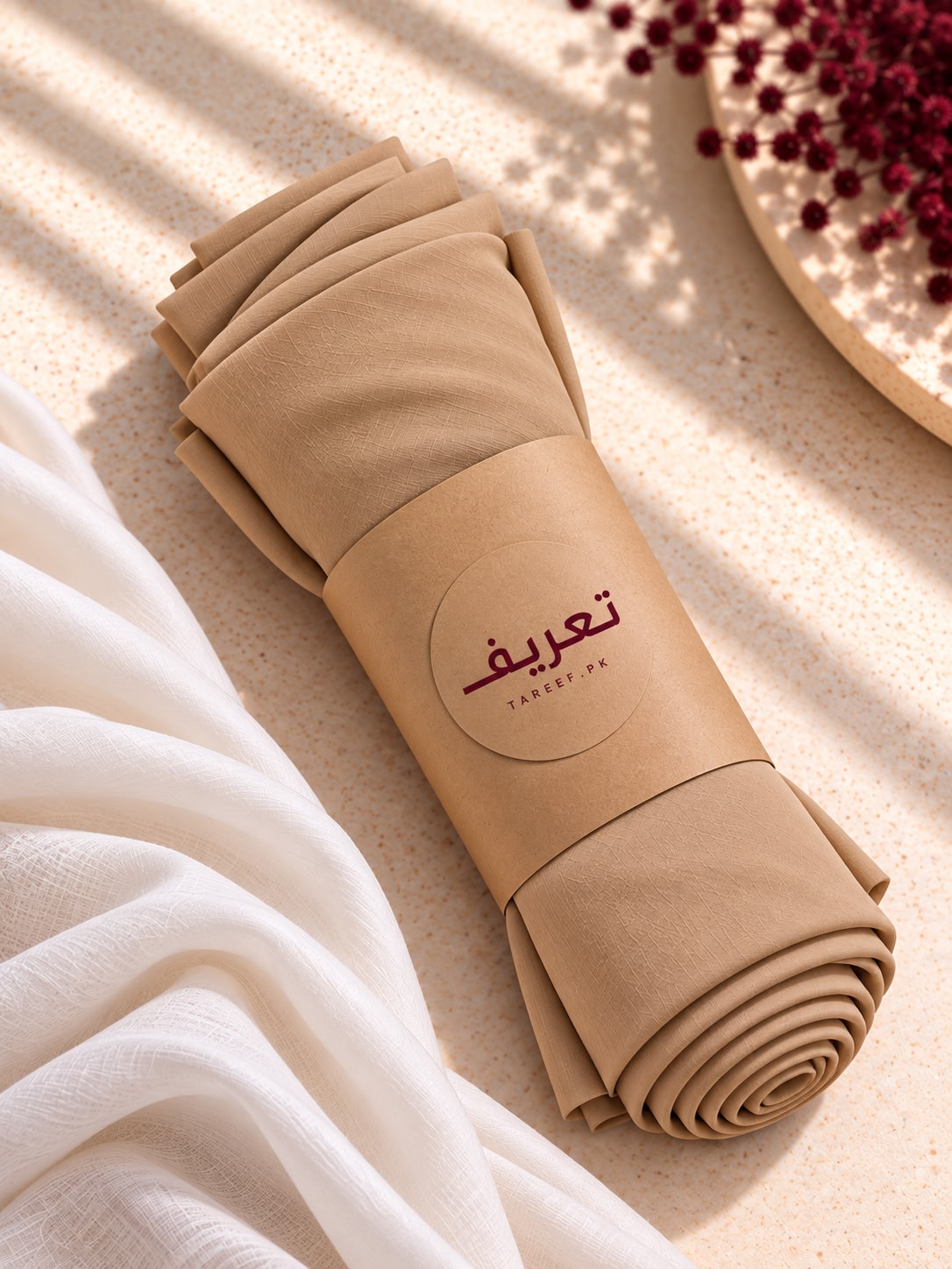 A beige draped hijab styled on a white mannequin wearing a white long-sleeve dress, set in a warm, sunlit interior with a vase of red flowers on the left.