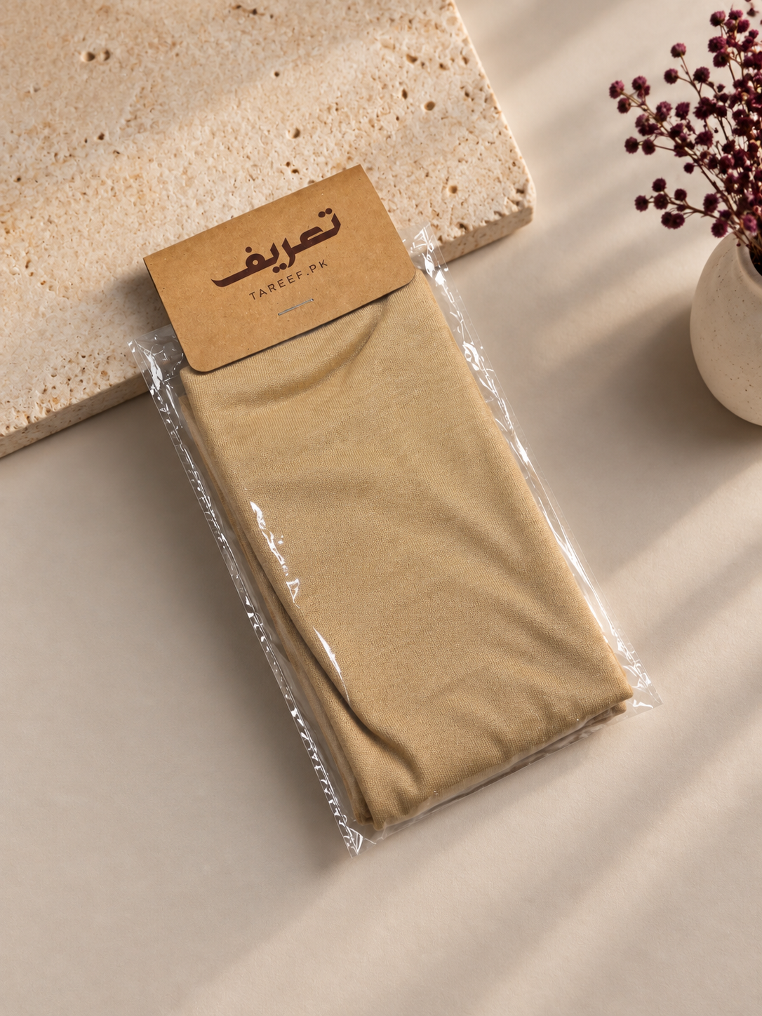 A folded beige hijab cap in clear plastic packaging with a tan paper label that reads 'TAREEF' styled on a beige surface with a stone slab and dried flowers in a vase.
