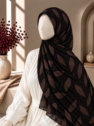 A chocolate black hijab with a brown leaf pattern and tiny white speckles, styled on a white mannequin dress in a warm, sunlit interior setting.