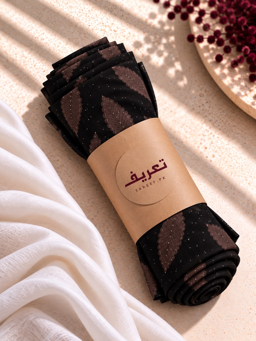A chocolate black hijab with a brown leaf pattern and tiny white speckles, styled on a white mannequin dress in a warm, sunlit interior setting.