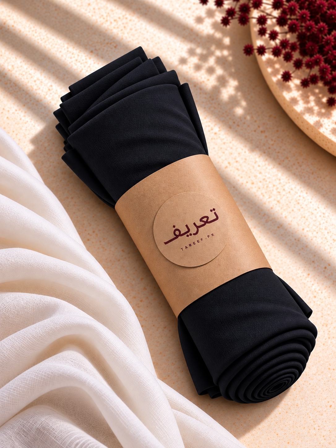 A black hijab wrapped in a kraft paper band with a circular logo 'TAREEF,' styled on a warm beige surface with draped white fabric and red dried flowers.