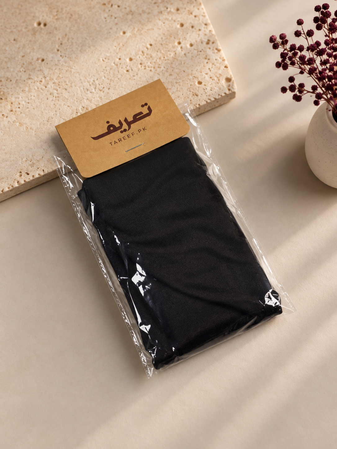 A folded black hijab cap packaged in a clear plastic sleeve with a kraft paper header card, placed on a beige surface with a stone slab and dried flowers in a vase nearby.
