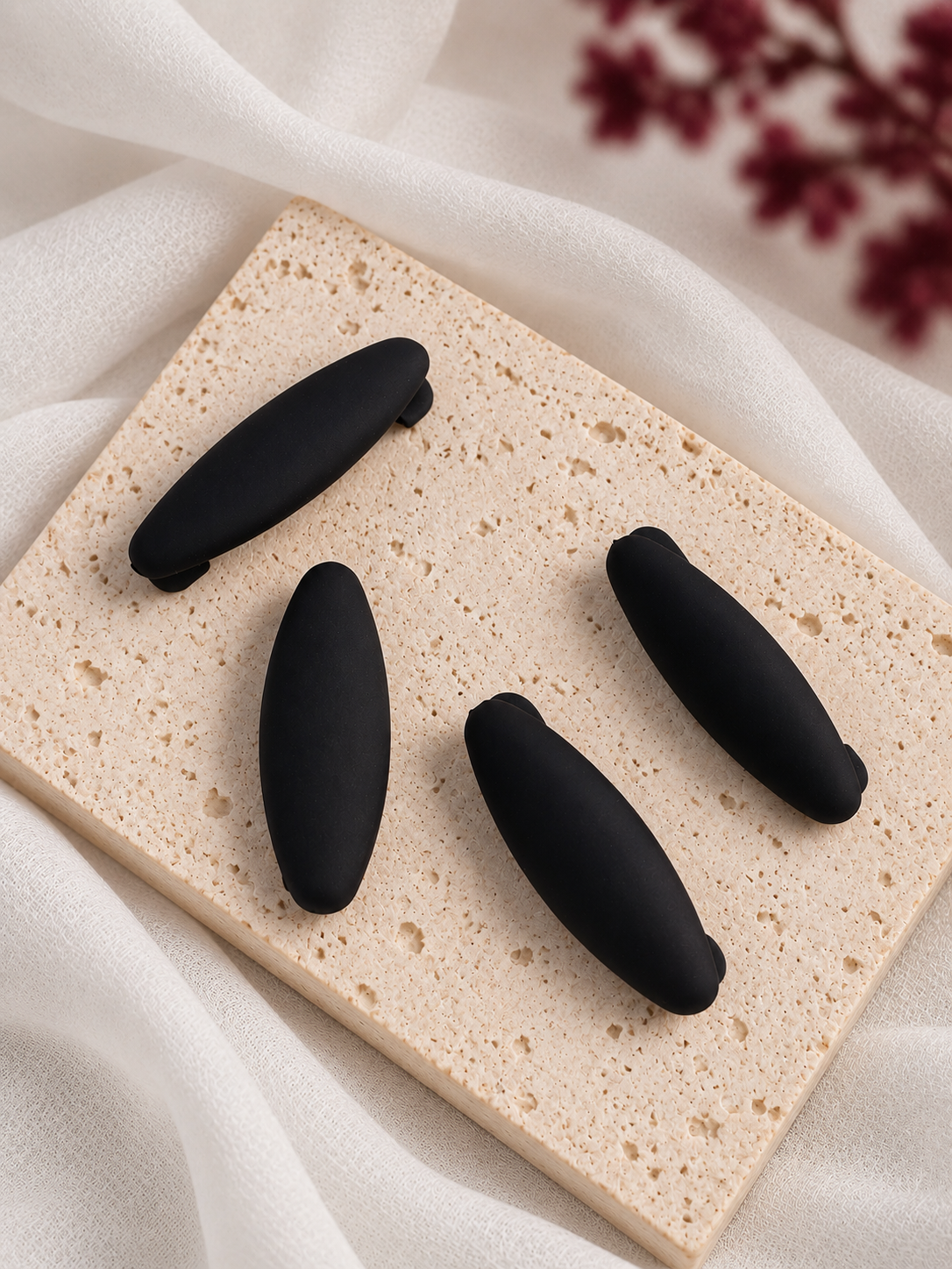 Four black matte hijab clips arranged on a rectangular beige stone slab with draped white fabric and blurred dark red flowers in the background.