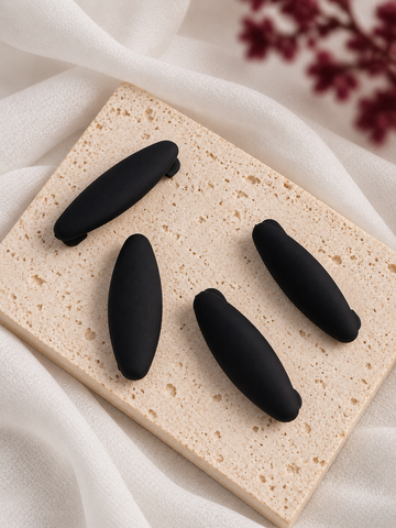 Four black matte hijab clips arranged on a rectangular beige stone slab with draped white fabric and blurred dark red flowers in the background.