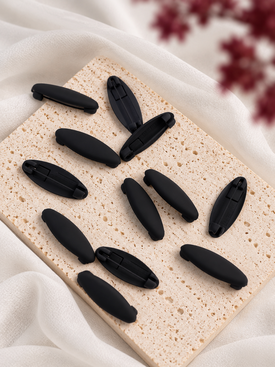 Twelve black matte hijab clips arranged on a textured beige stone slab with draped white fabric and blurred burgundy flowers in the background.