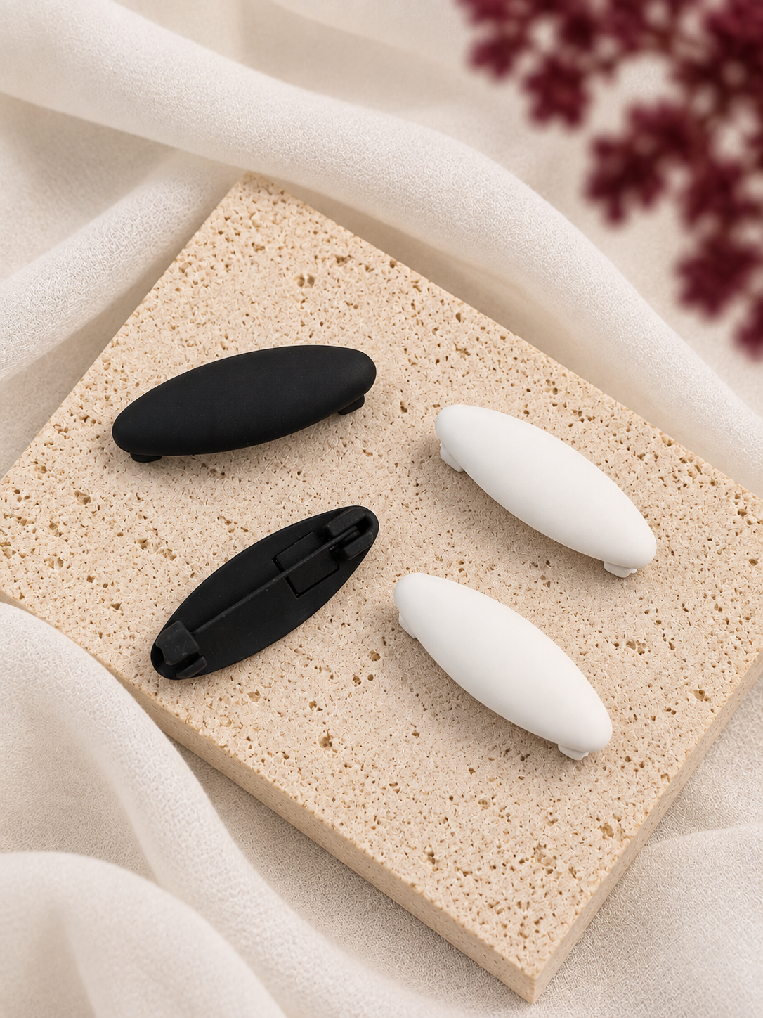 Four small matte hijab clips in two black and two white colors, arranged on a rectangular beige stone slab with draped cream fabric and blurred burgundy flowers in the background.