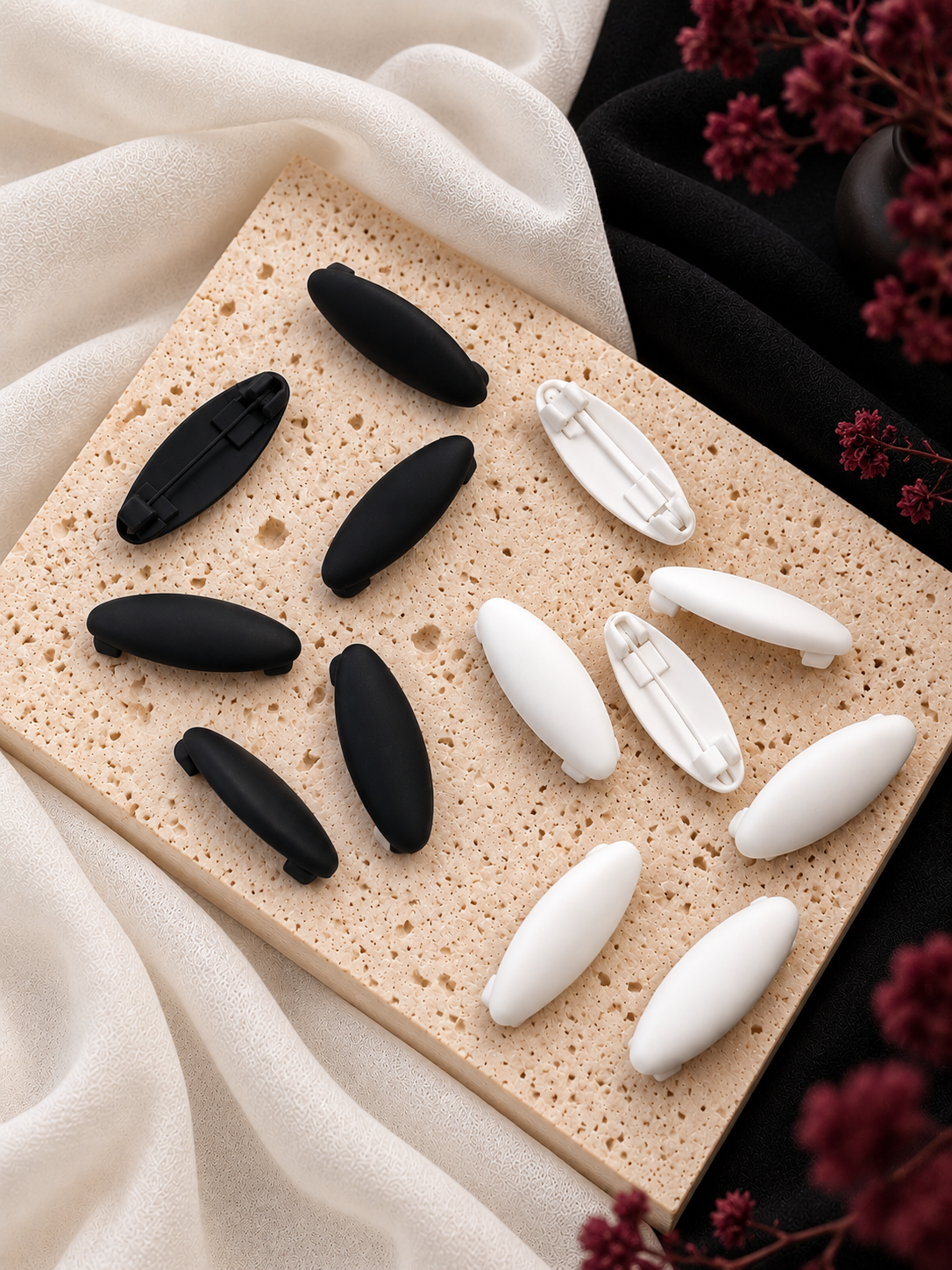 Four small matte hijab clips in two black and two white colors, arranged on a rectangular beige stone slab with draped cream fabric and blurred burgundy flowers in the background.