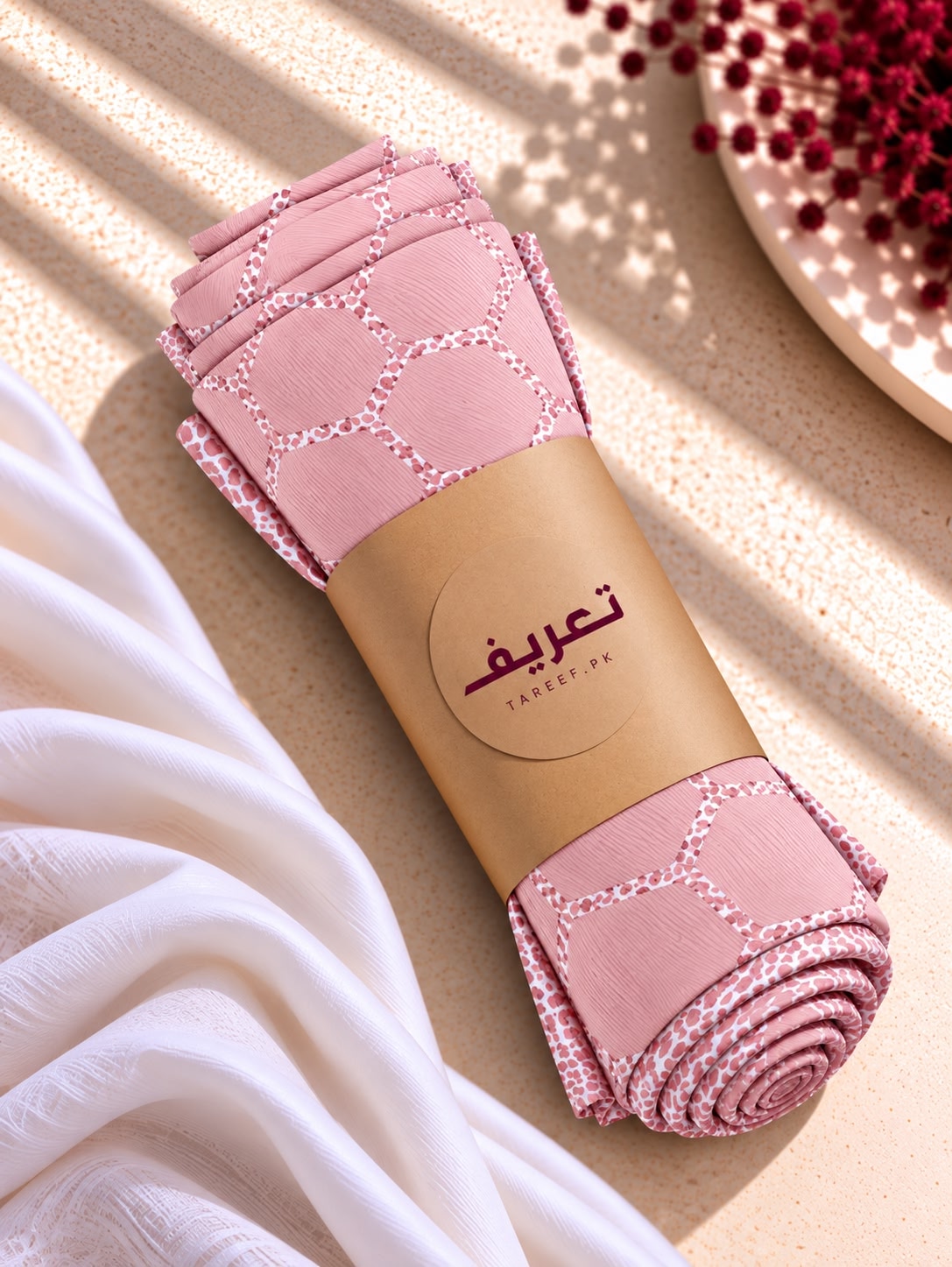 A pink embroidered hijab draped over a mannequin wearing a white long-sleeve top, set in a warm, sunlit interior with a vase of red flowers and neutral walls.