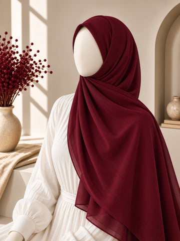 A deep burgundy hijab draped elegantly on a white mannequin wearing a white long-sleeve dress, set in a warm, sunlit interior with a vase of red berries in the background.