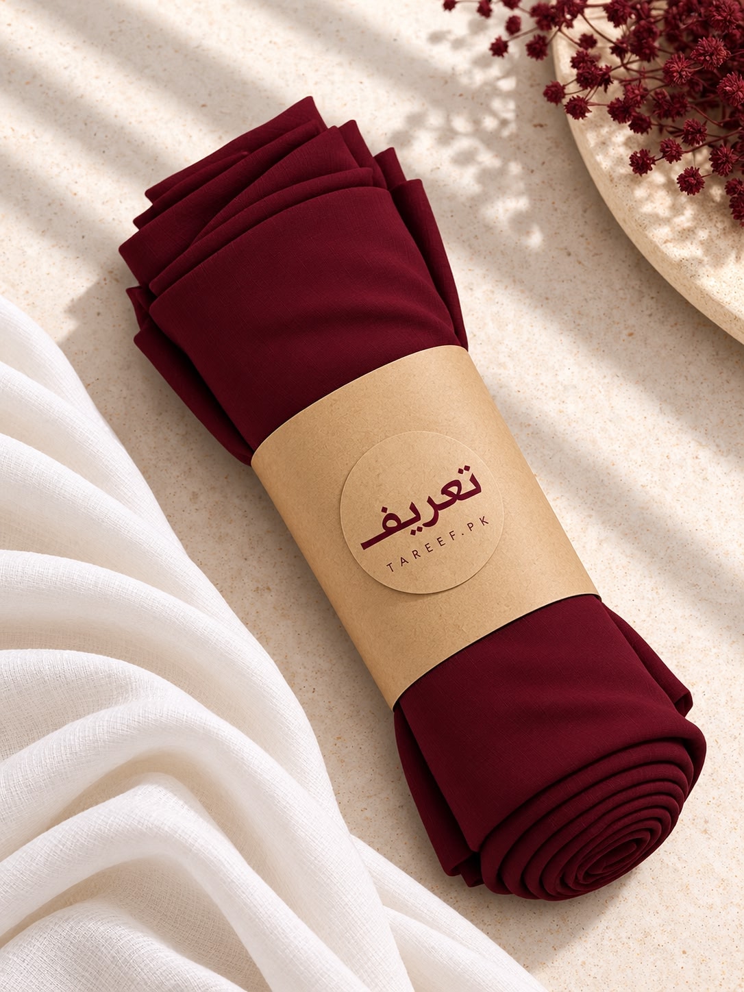 A folded burgundy hijab wrapped in a kraft paper band with the brand name 'TAREEF' styled on a light beige surface with draped white fabric and dried flowers.