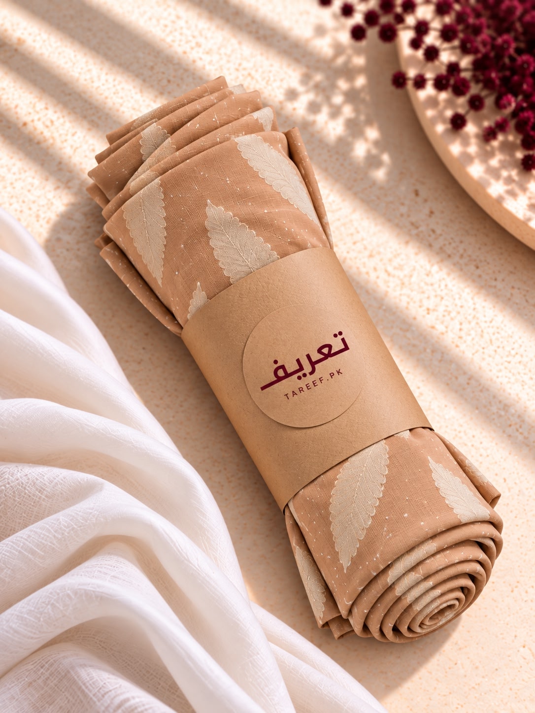 A draped hijab in a warm beige and cream leaf pattern, styled on a white mannequin wearing a cream dress in a warm, sunlit interior setting.