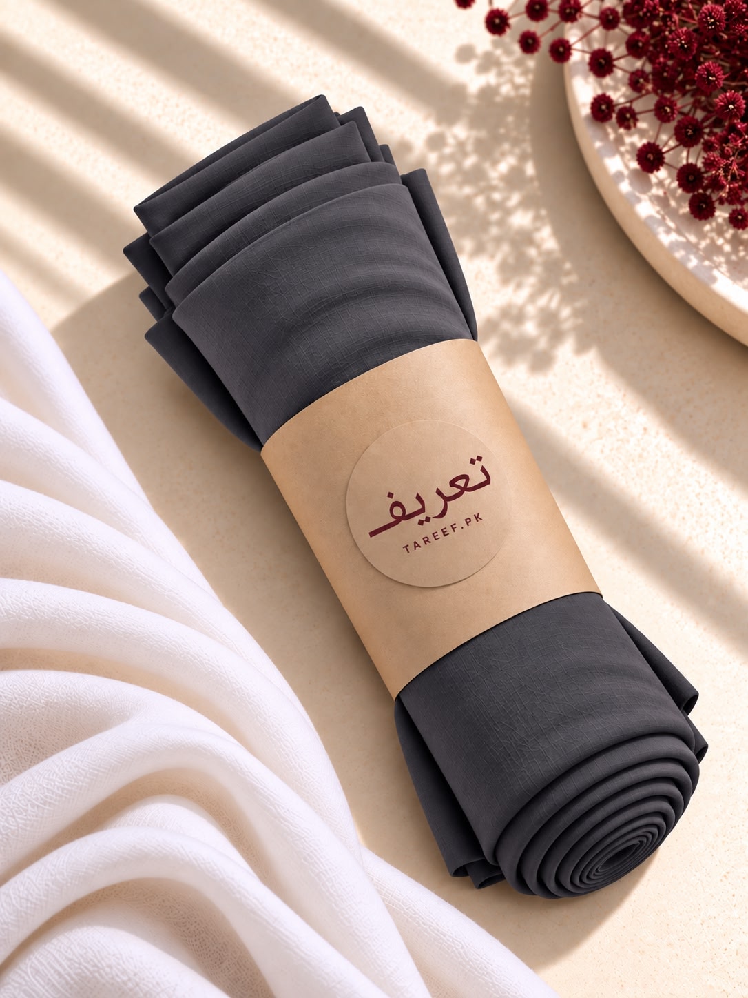 A dark hijab wrapped in a kraft paper band with brand name 'TAREEF' visible, styled on a beige surface with white draped fabric and red berries in the corner.
