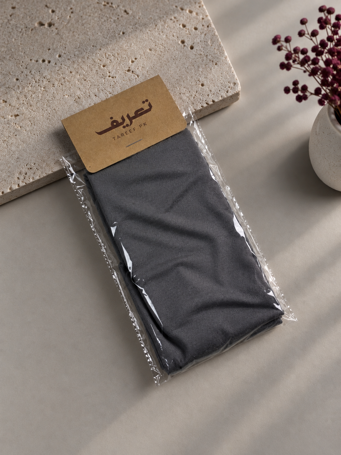 A dark gray hijab cap tied in a knot at the back, displayed on a mannequin head wearing a black top. The background is a warm beige wall with soft diagonal shadows, and a decorative vase with dried flowers is visible on the left.