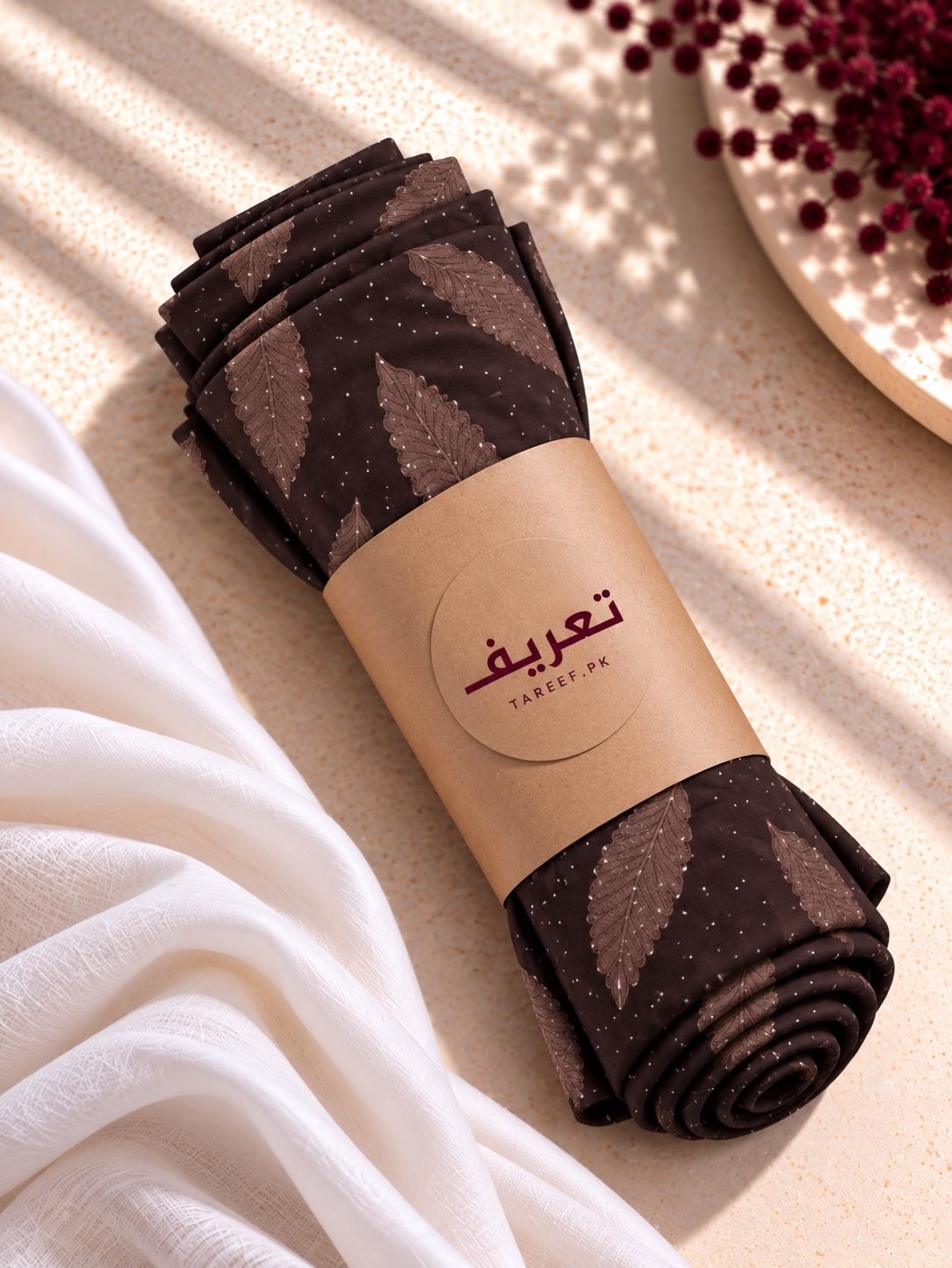A rolled of dark brown hijab with a kraft paper band labeled 'TAREEF' styled on a light beige surface with white fabric and red berries.