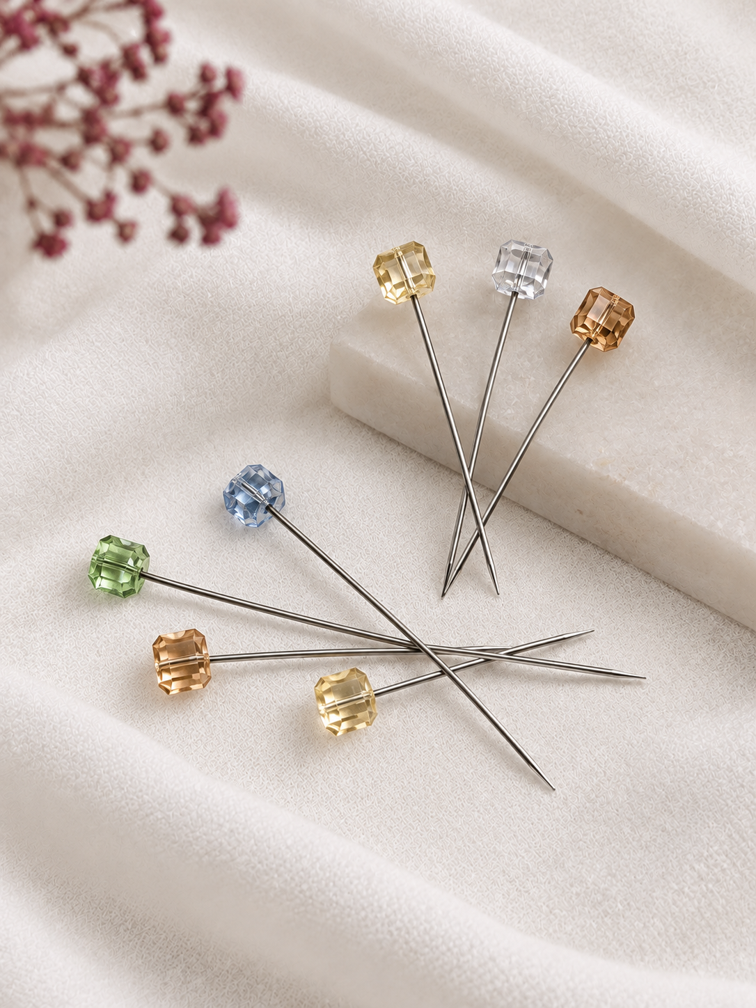 Eleven metal Crystal Hijab Pins arranged in a circular pattern, each topped with a small faceted gemstone in different colors, including blue, green, yellow, and clear, displayed on a soft white fabric background with dried flowers in the corners.