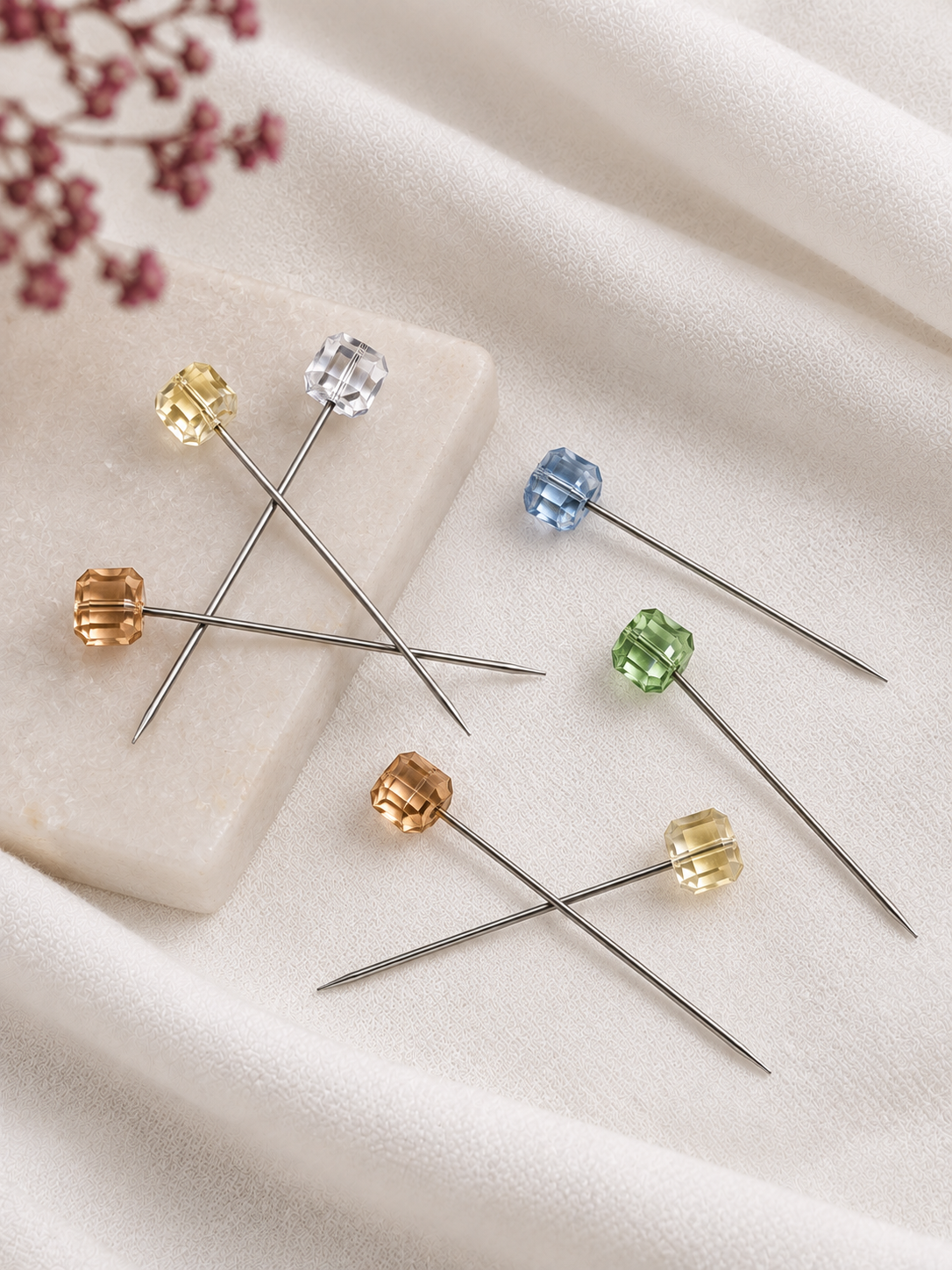 Seven Crystal Hijab Pins with faceted crystal tops in yellow, clear, blue, green, and rose gold, arranged on a soft cream fabric background with a small decorative floral sprig.