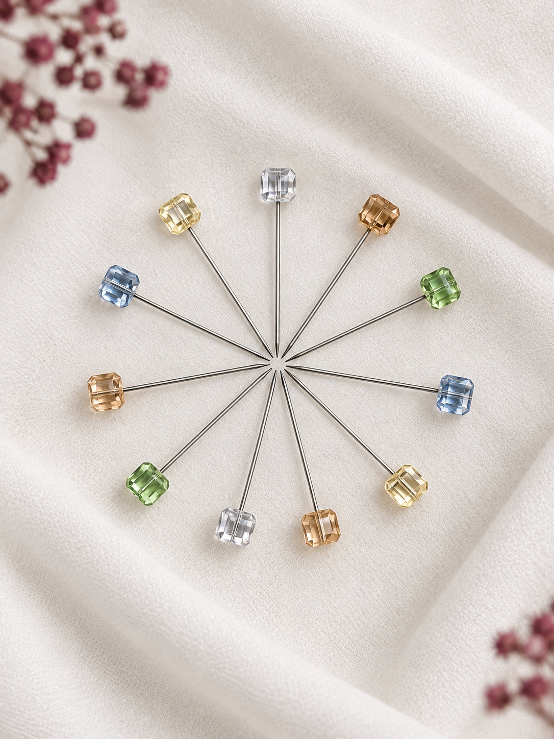 Eleven metal Crystal Hijab Pins arranged in a circular pattern, each topped with a small faceted gemstone in different colors, including blue, green, yellow, and clear, displayed on a soft white fabric background with dried flowers in the corners.