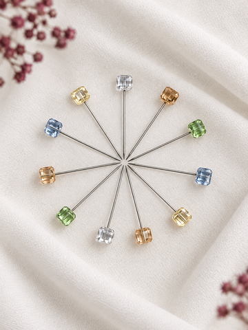 Eleven metal Crystal Hijab Pins arranged in a circular pattern, each topped with a small faceted gemstone in different colors, including blue, green, yellow, and clear, displayed on a soft white fabric background with dried flowers in the corners.
