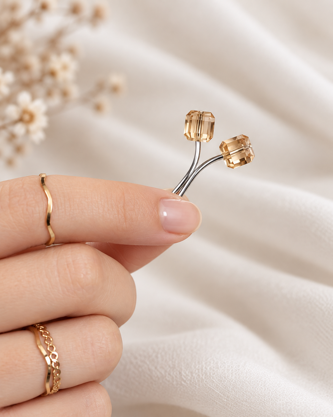 A close-up of a hand holding a curved hijab loop pin, against a soft cream fabric background with blurred dried flowers.