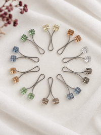 Ten curved hijab loop pins with translucent colored beads in various colors, arranged in a circular pattern on a soft white fabric background.