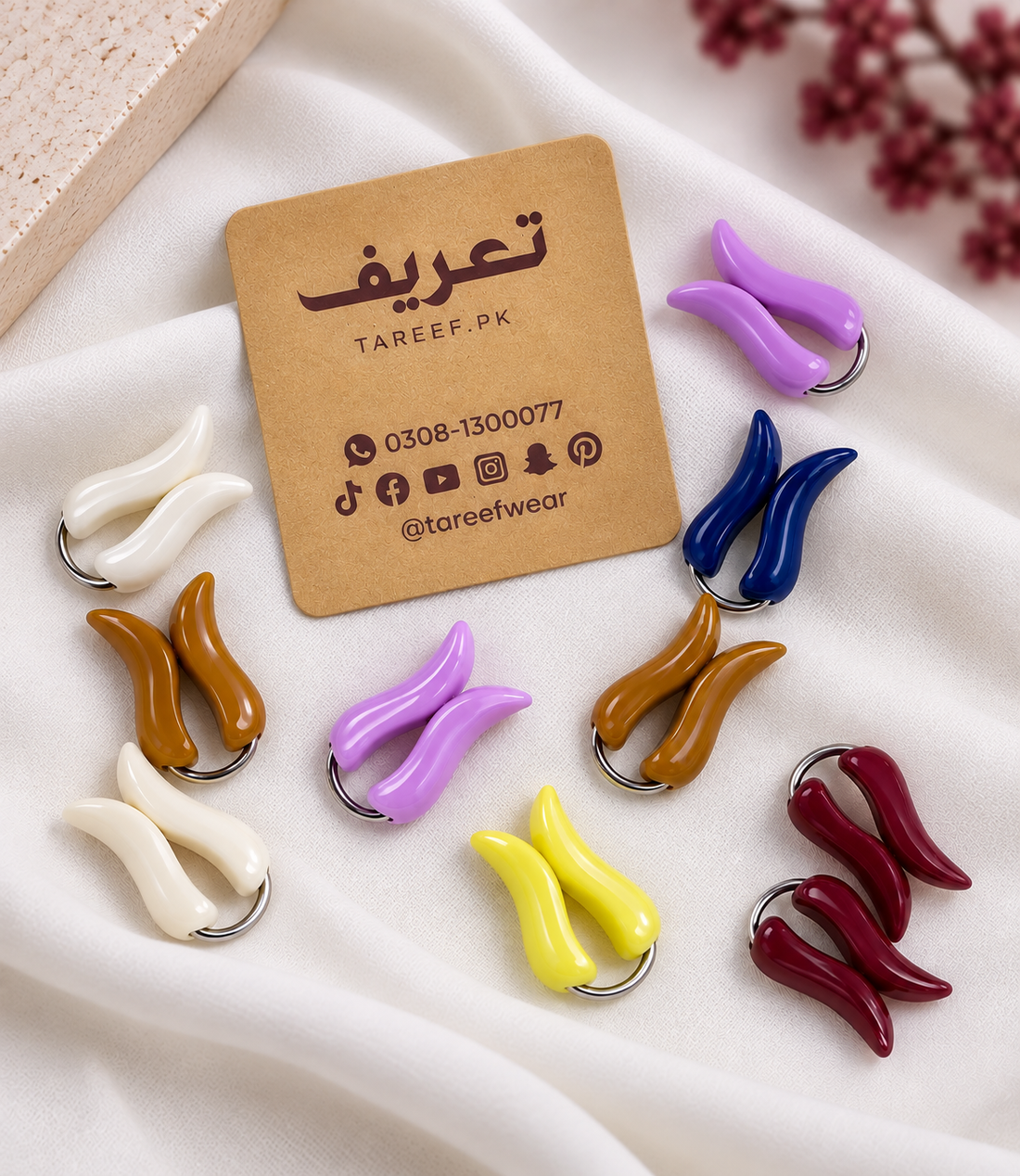 A circular arrangement of colorful curved hijab loop pins on a soft white fabric background with dried floral accents in the corners. The markers come in multiple colors, including navy blue, cream white, yellow, lavender purple, brown, burgundy, and peach, each with a small silver metal ring.