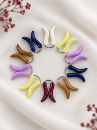 A circular arrangement of colorful curved hijab loop pins on a soft white fabric background with dried floral accents in the corners. The markers come in multiple colors, including navy blue, cream white, yellow, lavender purple, brown, burgundy, and peach, each with a small silver metal ring.
