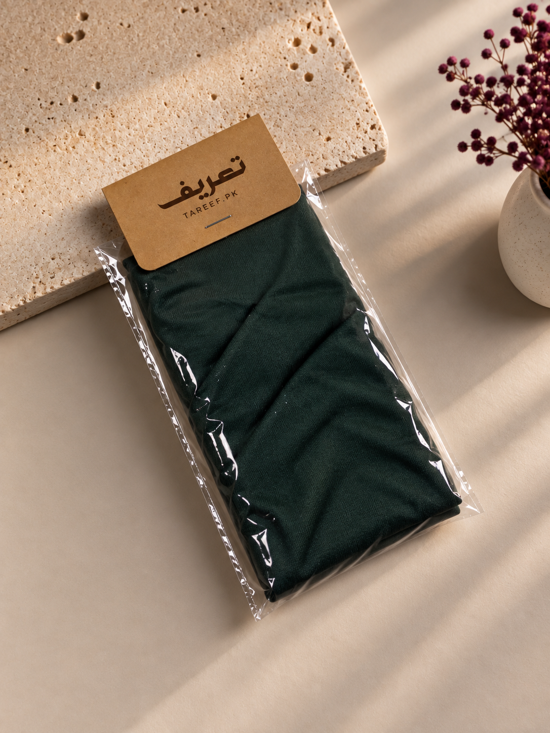 A dark green folded hijab cap sealed in a clear plastic bag with a kraft paper label reading 'TAREEF' styled on a beige surface with a stone slab and dried flowers in the background.
