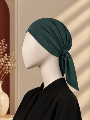 A dark teal-green hijab cap tied in a knot on a white mannequin head, styled with a black top against a warm beige background with soft window shadow patterns.