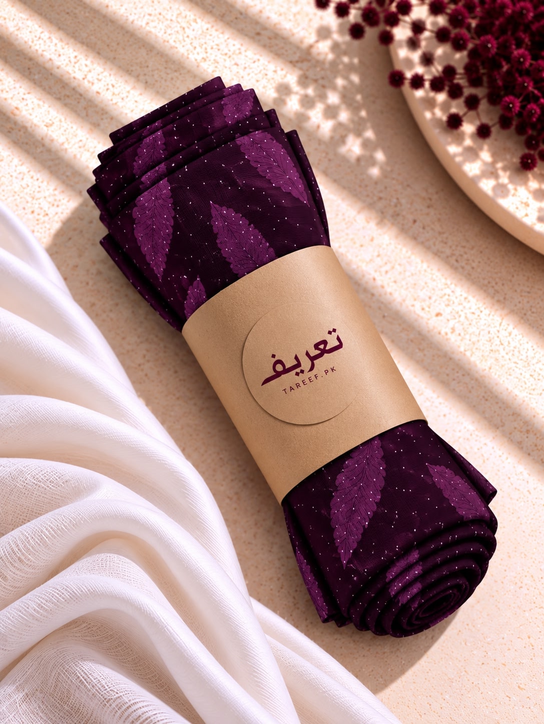 A dark purple hijab with a subtle leaf pattern and tiny gold glitter flecks, styled on a mannequin wearing a white dress in a warm, sunlit interior setting.