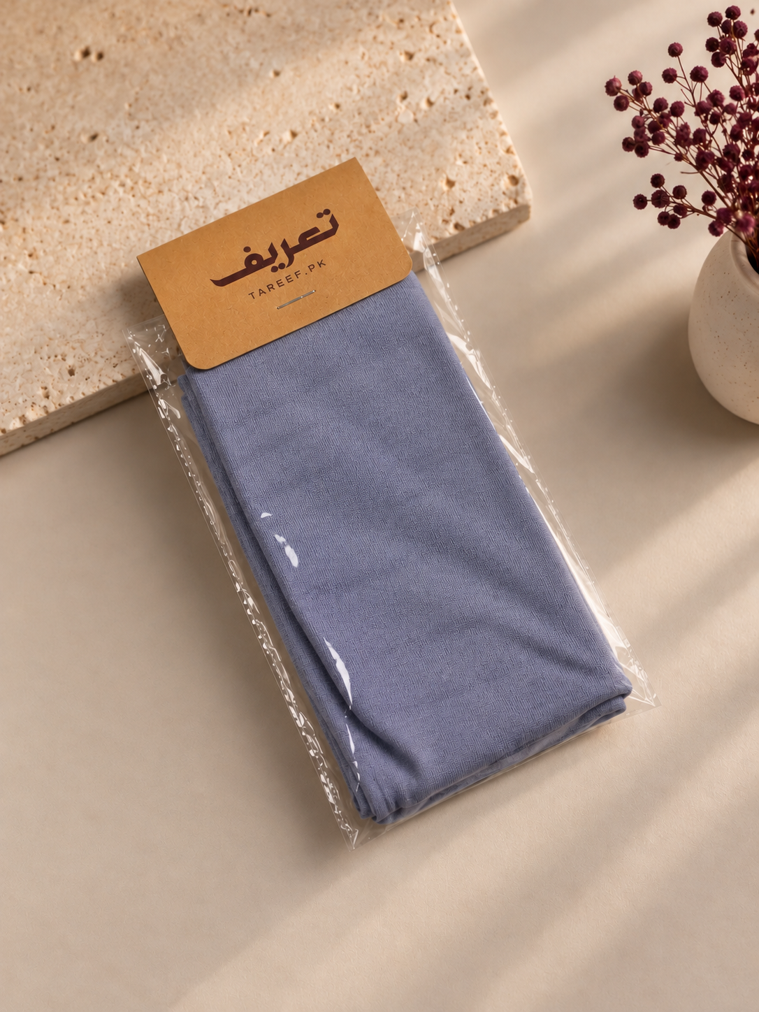 A slate blue hijab cap tied in a knot on a mannequin head, styled with a black top against a warm beige background with soft window shadows.