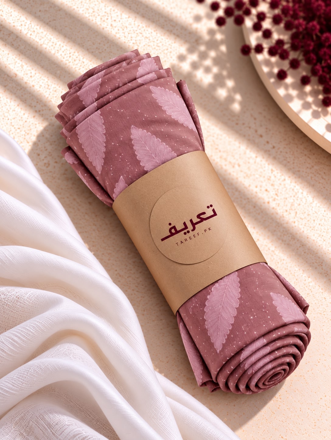 A dusty pink hijab with a lighter pink leaf pattern draped over a white dress on a mannequin, set in a warm, sunlit interior with a vase of red berries in the background.