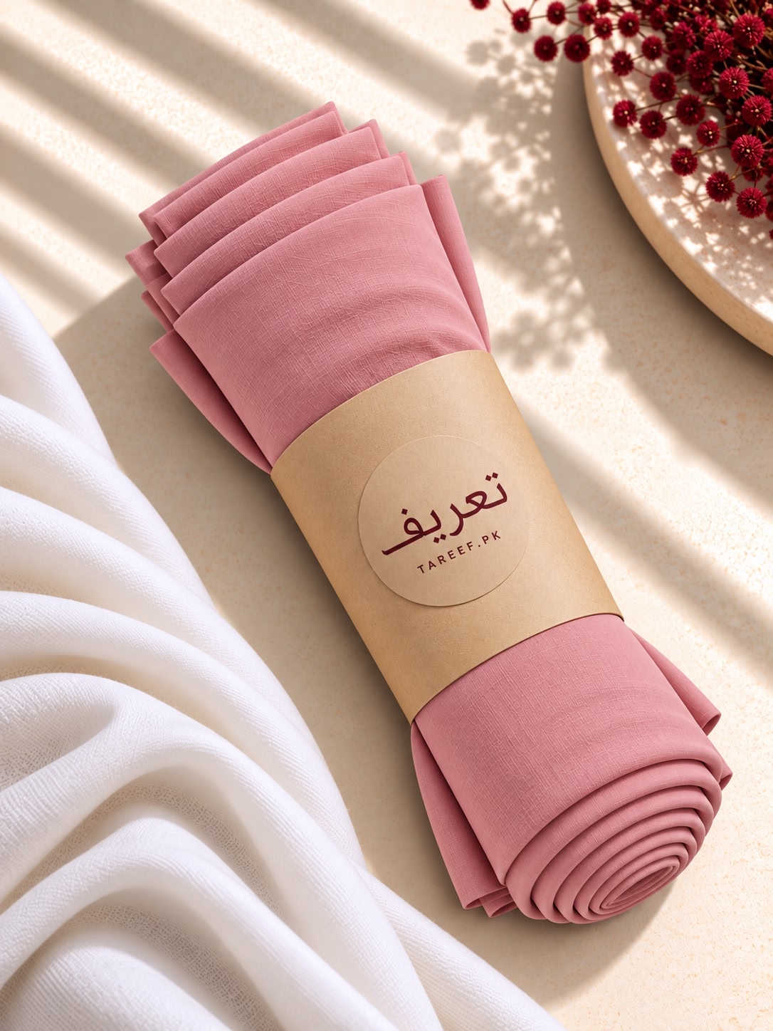 A rolled dusty pink hijab wrapped in a kraft paper band with the brand name 'TAREEF' styled on a beige surface with a white draped cloth and red berries in the background.