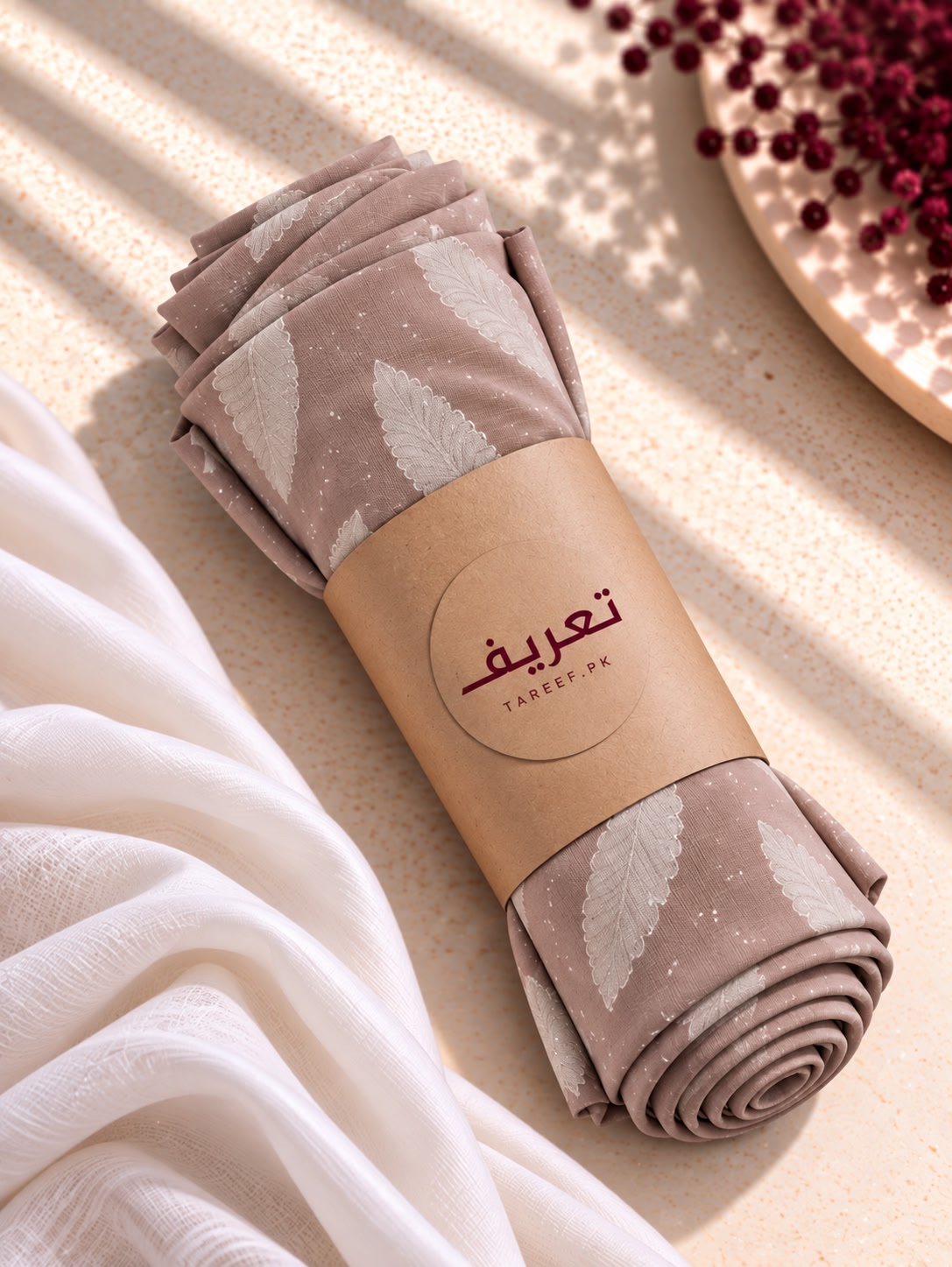 A rolled hijab in a dusty taupe color with a white leaf pattern, wrapped in a kraft paper band with the brand name 'TAREEF'. The product is styled on a textured beige surface with soft white draped fabric and shadows.