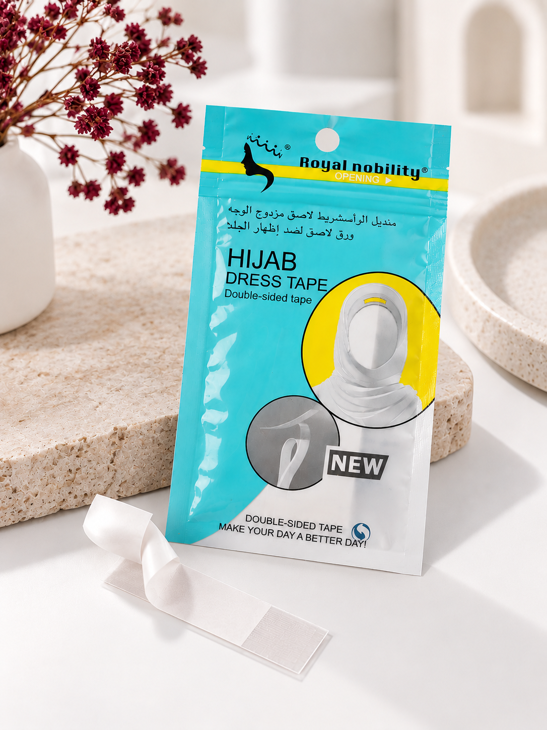 A package of Royal Nobility Organics Hijab Double Tape is displayed on a white surface with a small piece of the white double-sided tape in front. The background features a beige stone slab with dried red flowers in a white vase and a ceramic dish.