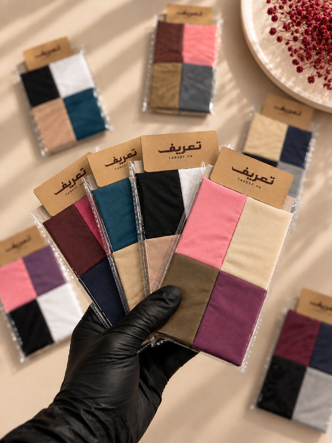 A hand wearing a black glove holds packaged hijab tubes. The hijab tubes include pink, beige, brown, navy, gray, and purple tones, arranged on a warm beige surface with other matching packs scattered around.