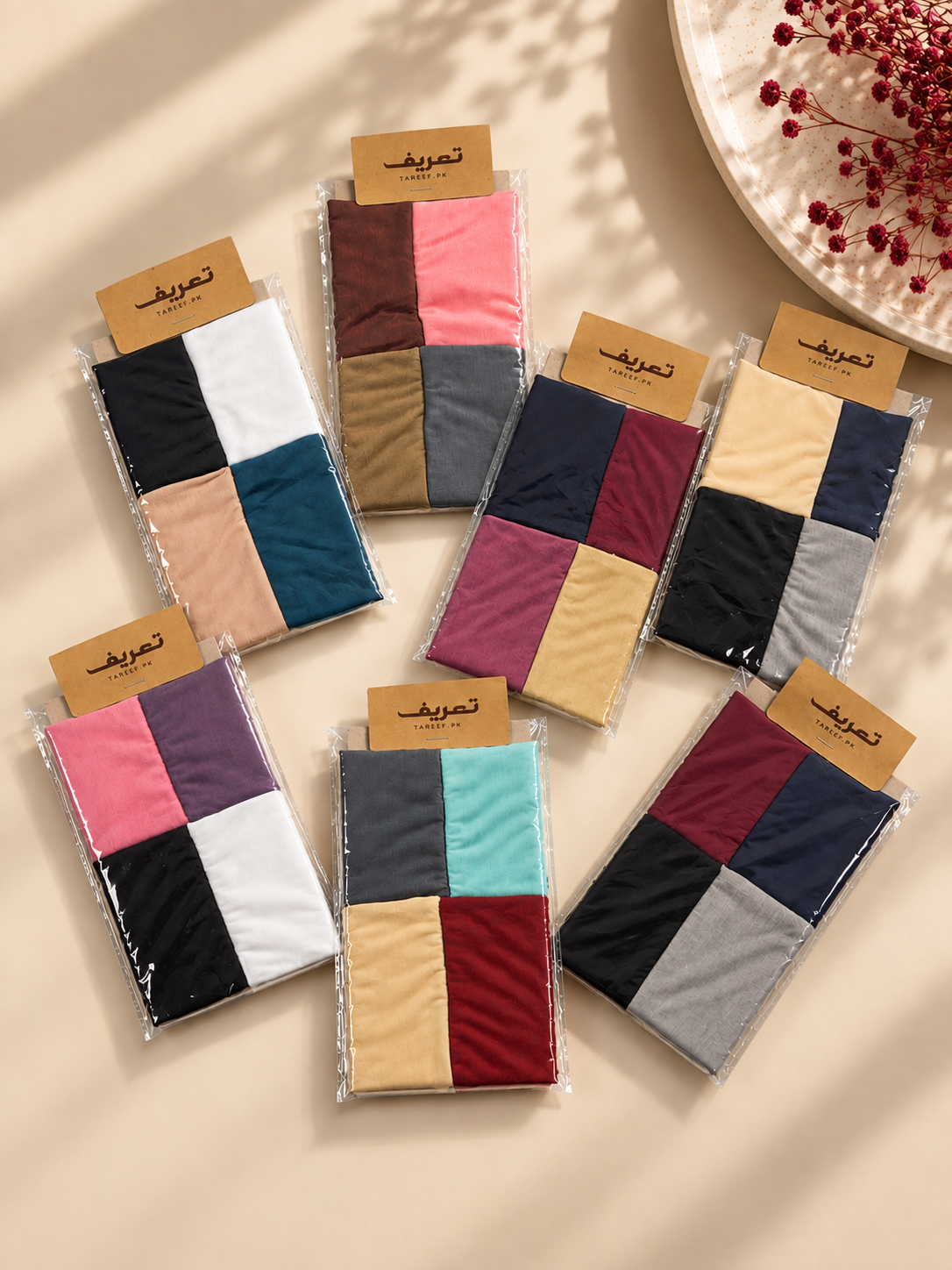 Seven packaged hijab tubes are arranged on a beige surface with decorative shadows and a plate of red berries in the corner. Each hijab tube shows four color blocks and has a small tan label 'TAREEF'.