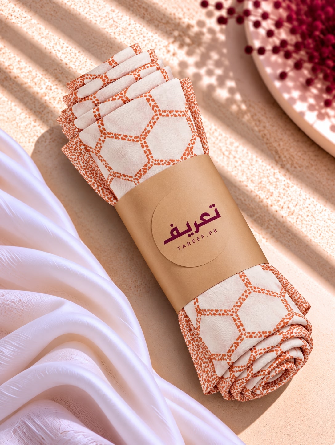 A white hijab with a rust-orange geometric lattice pattern and matching border, styled on a mannequin wearing a white dress in a warm, sunlit interior setting.
