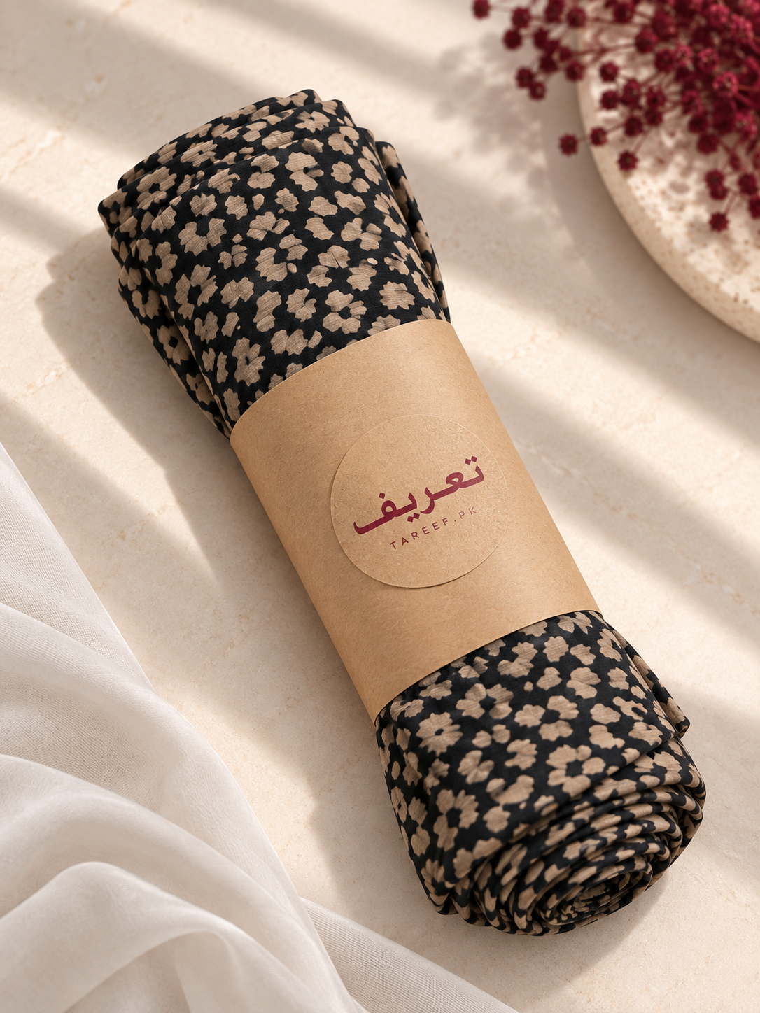 A rolled hijab wrapped in a brown paper band with the brand name 'TAREEF' set on a textured cream surface with soft white drapery and dried red flowers in the background.