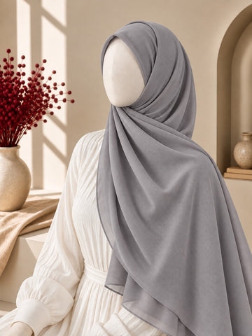 A light gray hijab draped elegantly on a mannequin wearing a white long-sleeve dress, set in a warm, sunlit interior with beige walls, a window frame, and a vase of red berries.