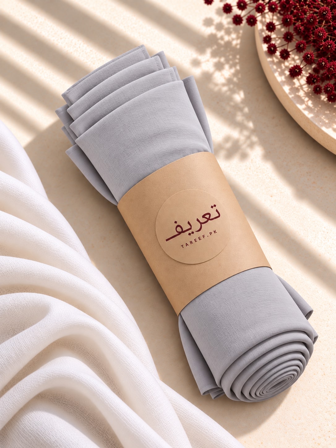 A rolled light gray hijab wrapped in a tan paper band 'TAREEF' visible, styled on a warm beige surface with soft shadows and a white draped cloth nearby.