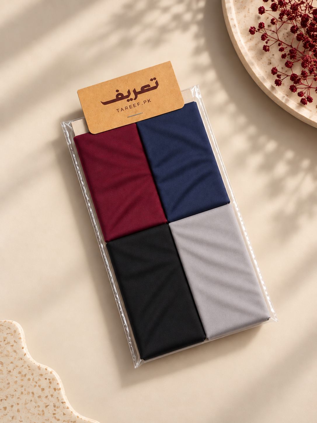 A packaged hijab tube in four colors: burgundy, navy, black, and light gray, with a kraft paper tag reading 'TAREEF' placed on top, photographed on a warm beige surface with decorative dried red flowers and soft shadows.