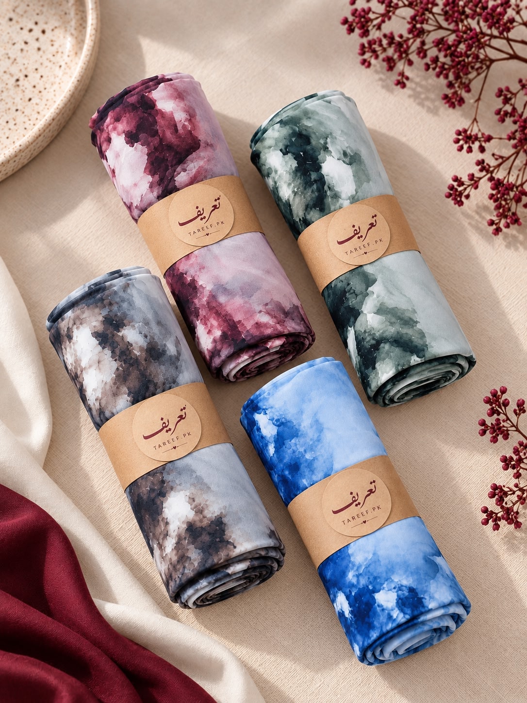 Four rolled hijabs with watercolor-style prints in pink, green, blue, and gray, each tied with a tan paper band featuring a circular logo. They are arranged on a beige fabric surface with decorative dried stems and draped red fabric.