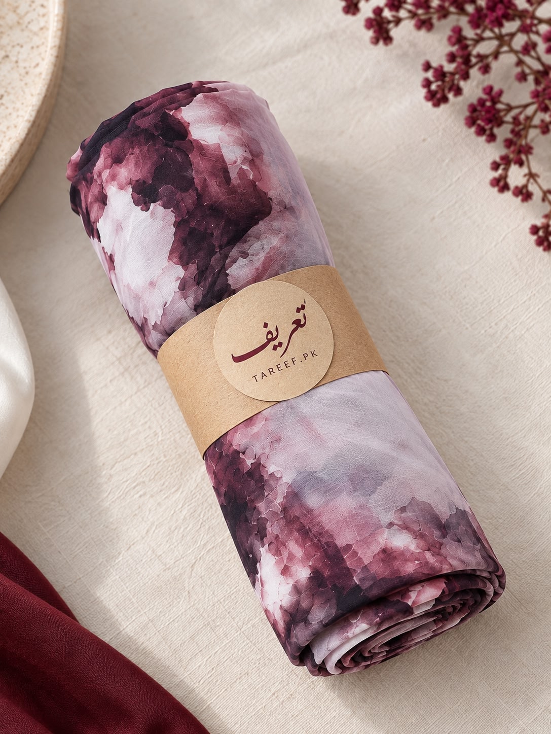A rolled hijab with a watercolor-style floral pattern in dusty pink, mauve, and white, wrapped with a kraft paper band labeled 'TAREEF', styled on a beige fabric surface with dried flowers and a small red cloth in the corners.