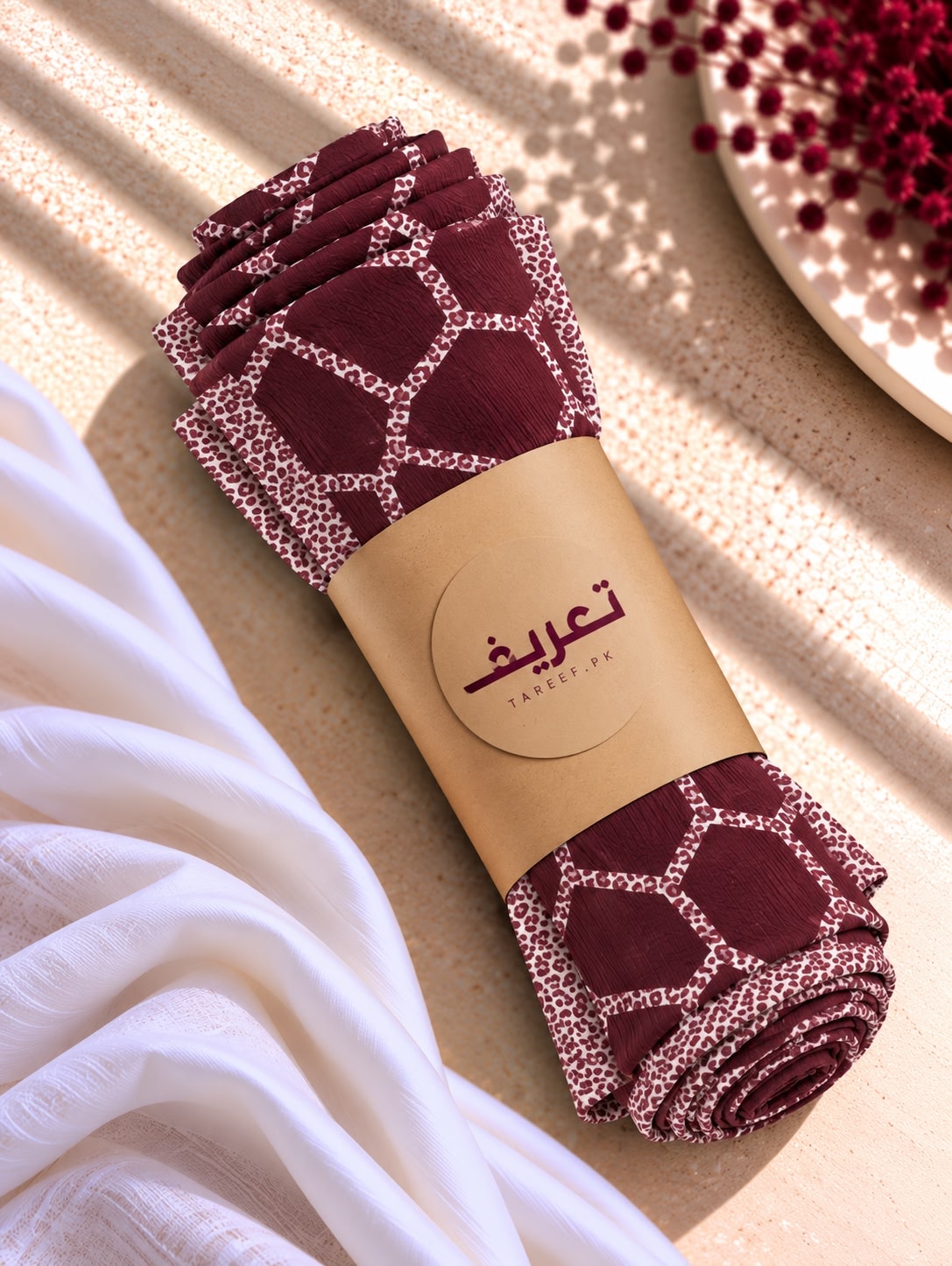 A rolled maroon and white patterned hijab wrapped in a tan paper band with Arabic text and the brand name 'TAREEF,' styled on a beige textured surface with draped white fabric and red floral decor.