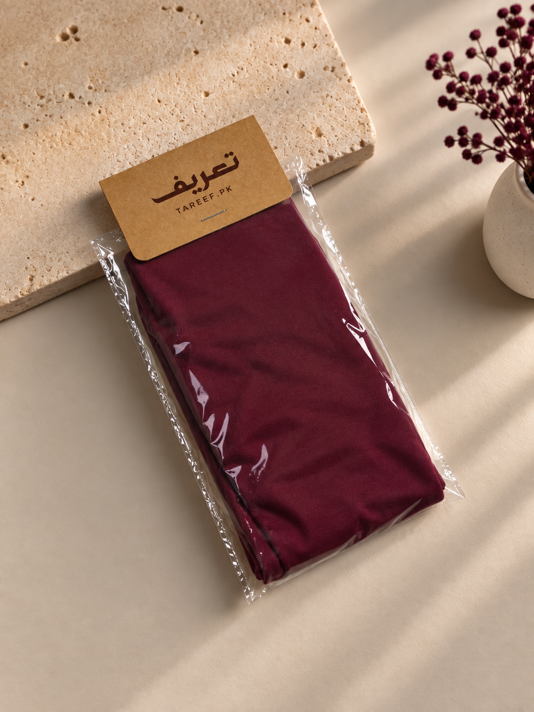 A folded maroon hijab cap in clear plastic packaging with a kraft paper labeled 'TAREEF' styled on a beige surface with a stone slab and dried flowers in the background.