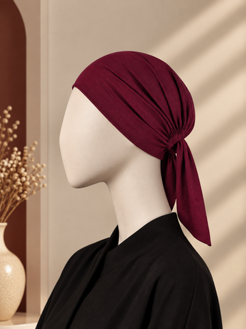 A deep burgundy knotted hijab cap displayed on a white mannequin head, styled with a black top against a warm beige background with soft window shadow patterns.