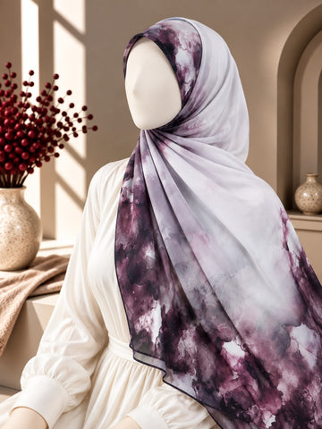 A lightweight hijab with a purple and white watercolor-like print, styled on a white mannequin wearing a white dress in a warm, sunlit interior setting.